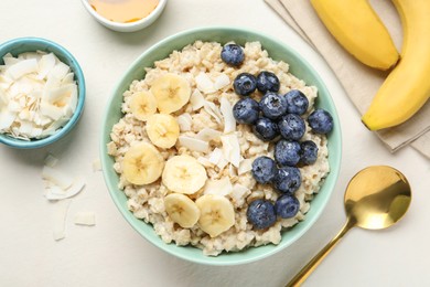 Tasty oatmeal with banana, blueberries, coconut flakes and honey served in bowl on beige table, flat lay