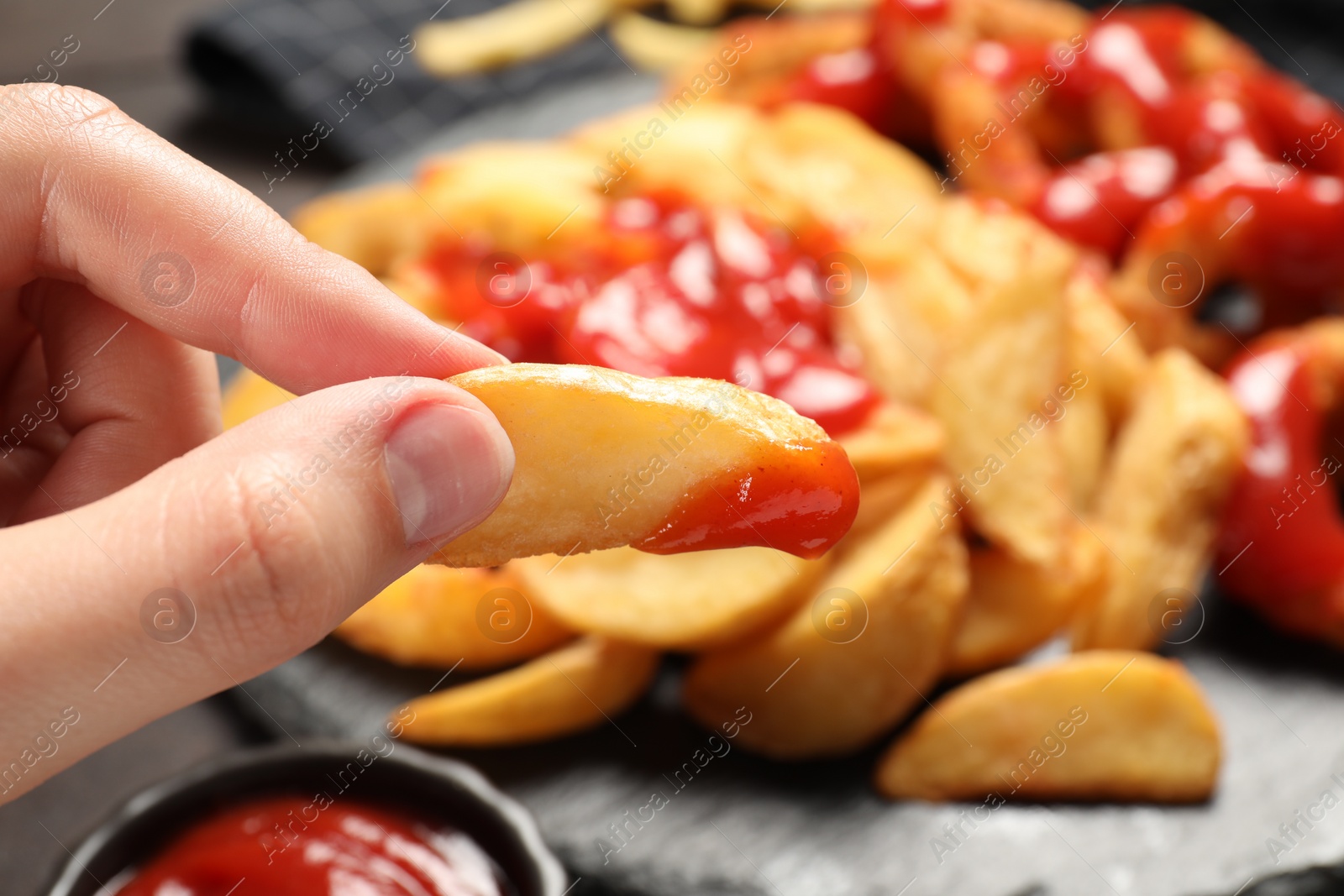 Photo of Woman holding delicious baked potato wedge with ketchup near different snacks at table, closeup
