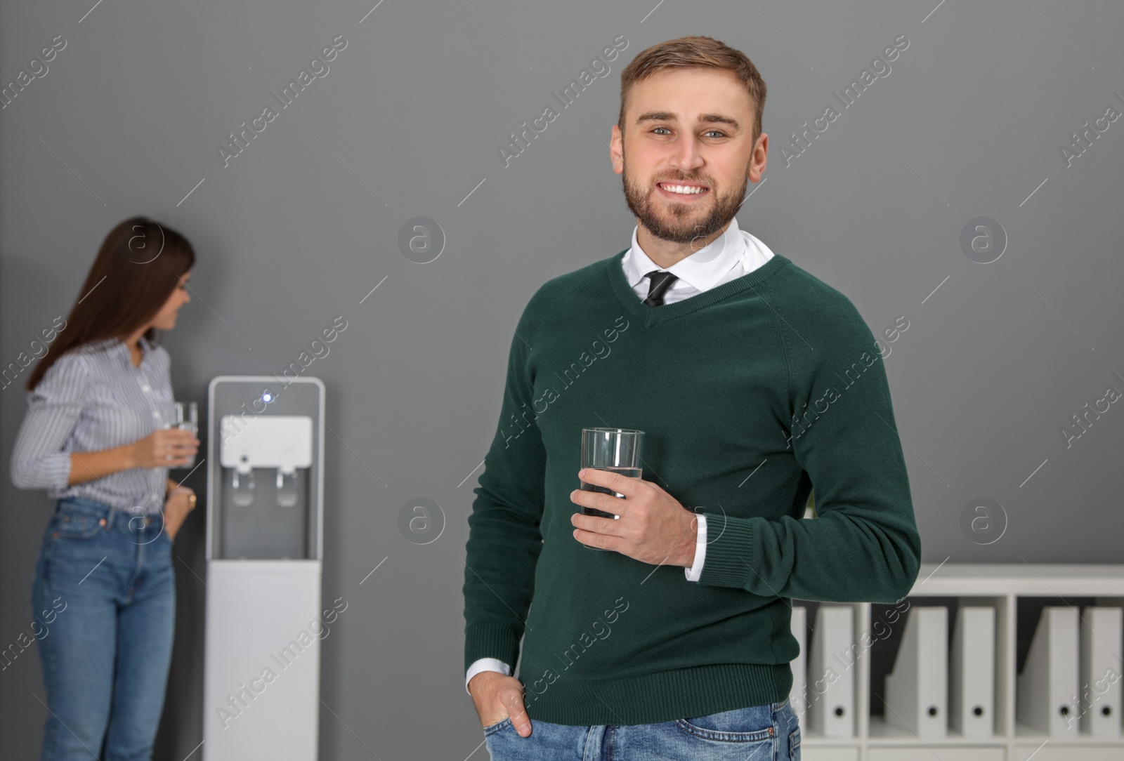 Photo of Young man with glass of water near cooler in office