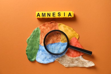 Word Amnesia made of yellow cubes, magnifying glass and brain with sections on orange background, flat lay