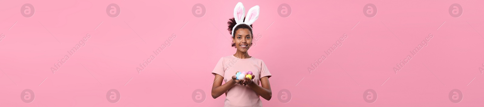 Image of African American woman with bunny ears holding Easter eggs on pink background. Banner design