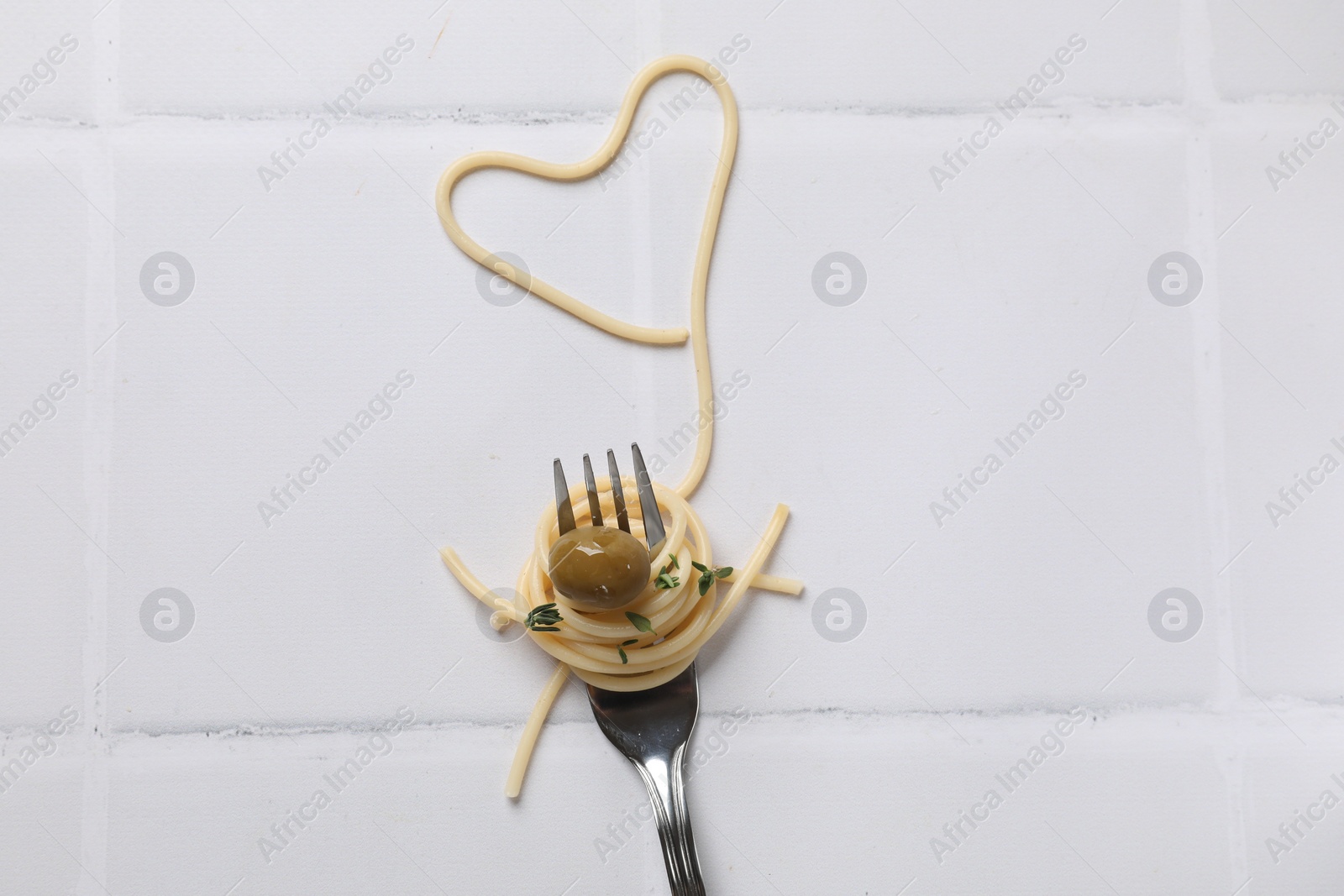 Photo of Heart made of tasty spaghetti, fork and olive on white tiled table, top view