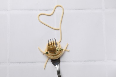 Heart made of tasty spaghetti, fork and olive on white tiled table, top view