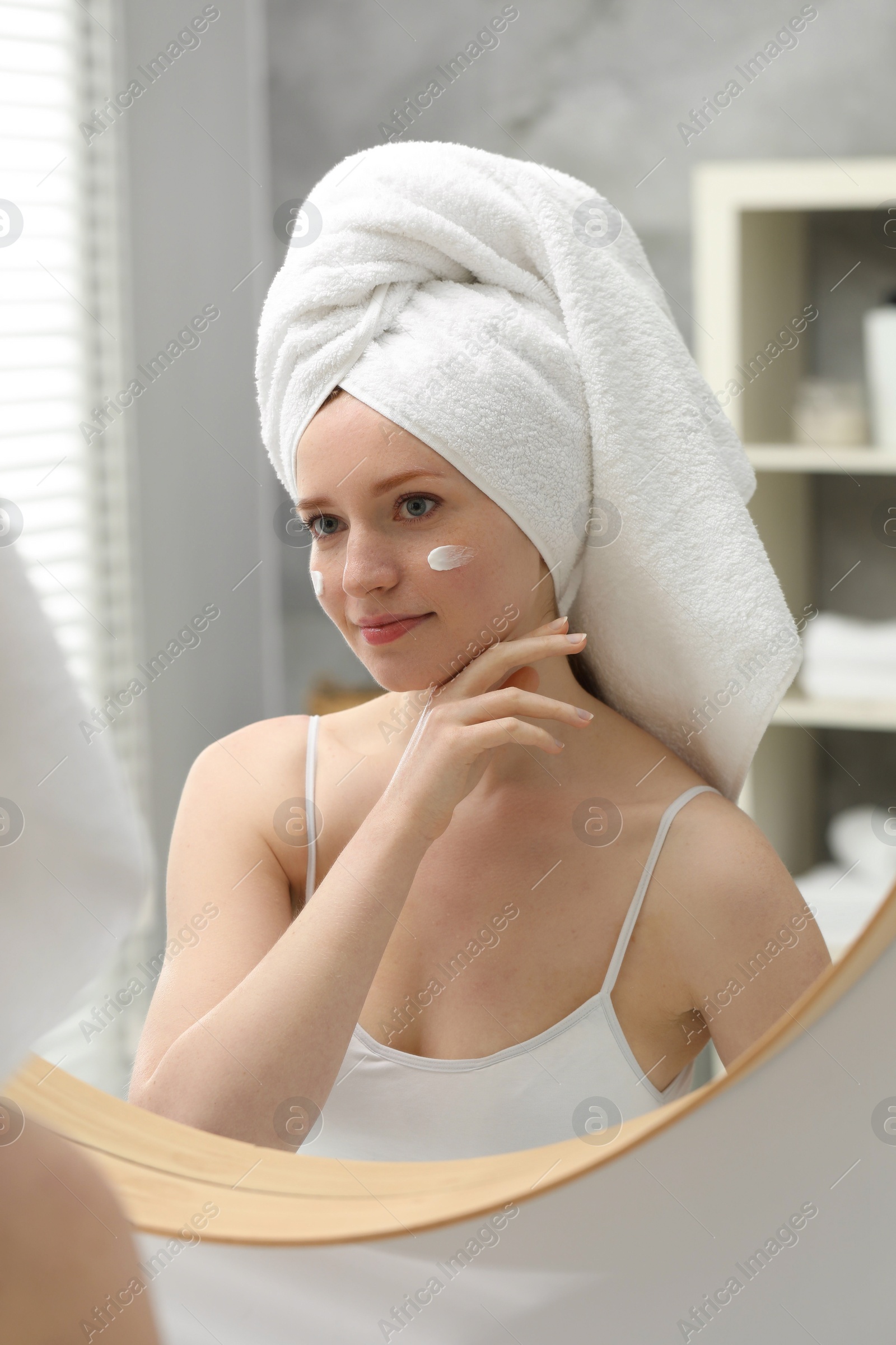 Photo of Beautiful woman with freckles and cream on her face near mirror in bathroom
