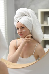 Beautiful woman with freckles and cream on her face near mirror in bathroom