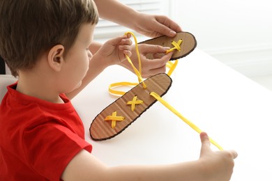 Mother teaching son to tie shoe laces using training cardboard template at white table, closeup