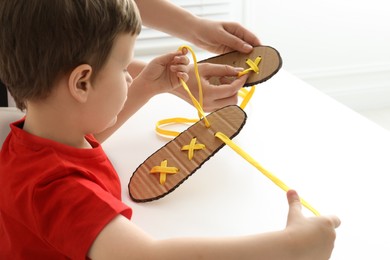 Photo of Mother teaching son to tie shoe laces using training cardboard template at white table, closeup