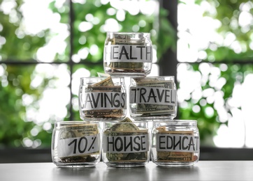 Photo of Glass jars with money for different needs on table against blurred background