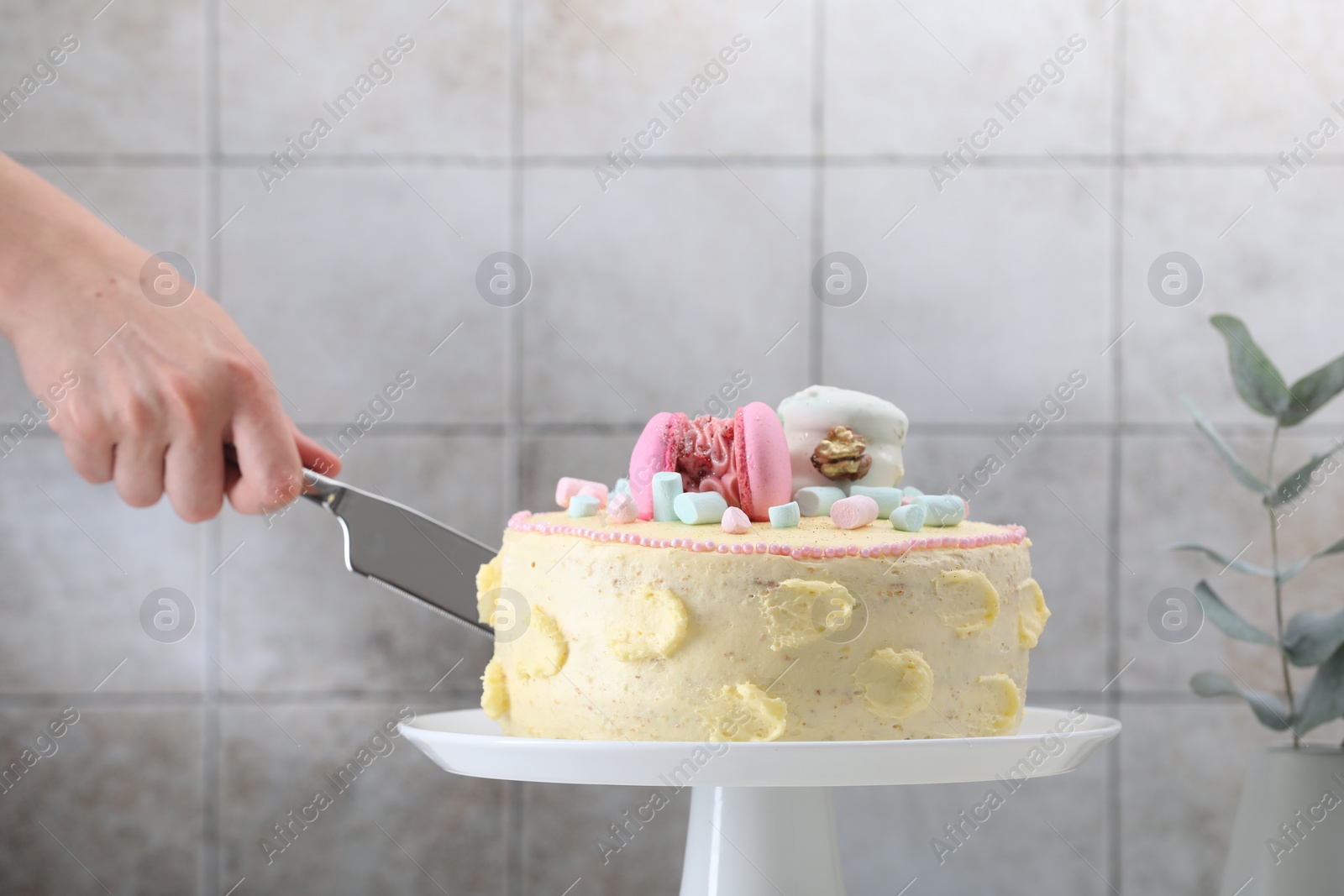 Photo of Woman cutting delicious cake decorated with macarons and marshmallows against light tiled background, closeup