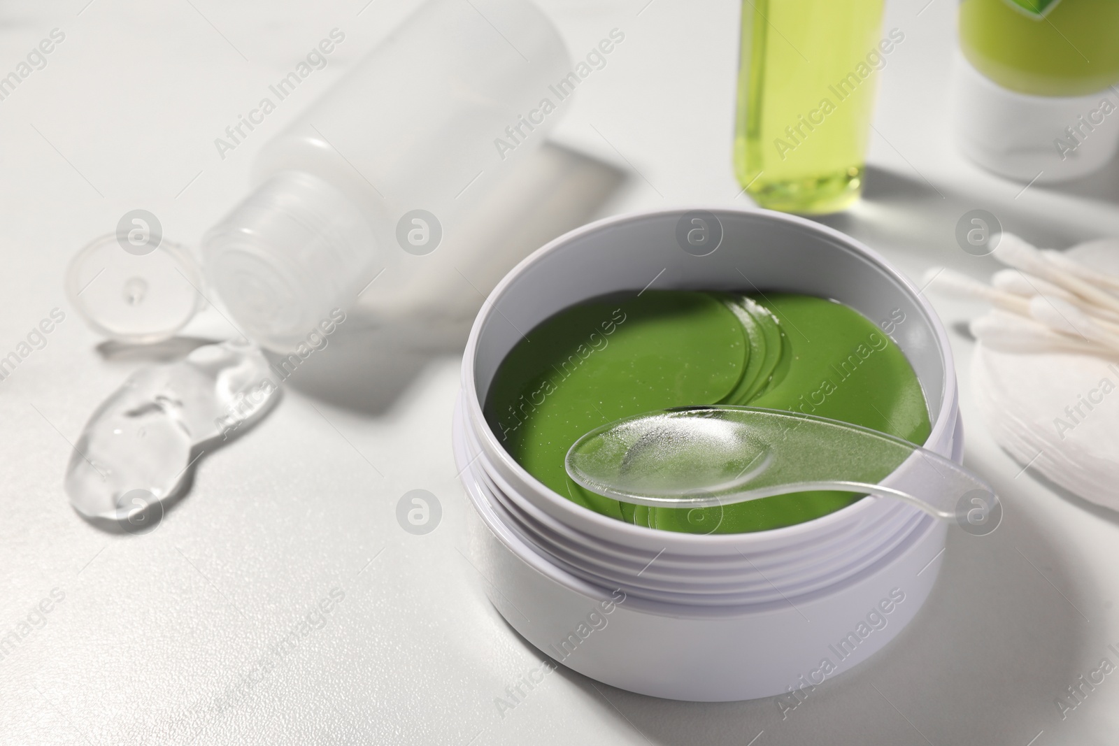 Photo of Jar of under eye patches with spoon and cosmetic products on white table, closeup