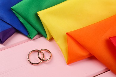 Rainbow LGBT flag and wedding rings on pink wooden table, closeup