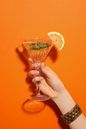 Woman holding martini glass of refreshing cocktail with lemon slice and rosemary on orange background, closeup