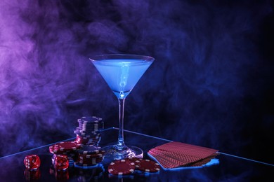 Photo of Casino chips, dice, playing cards and cocktail on dark background with smoke, space for text