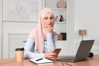 Muslim woman using smartphone near laptop at wooden table in room