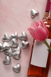 Delicious heart shaped chocolate candies, beautiful tulip and bottle of wine on pink table, flat lay