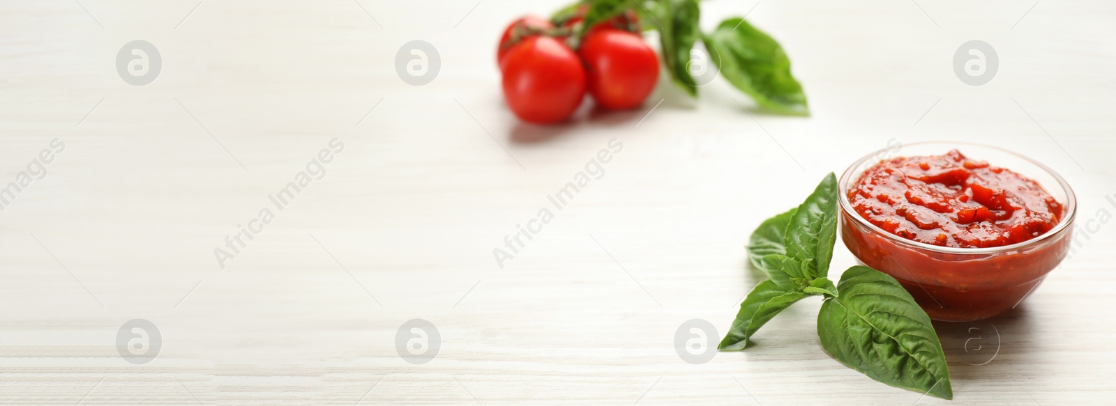 Photo of Delicious adjika sauce in glass bowl with basil on white wooden table. Space for text