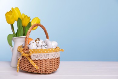 Wicker basket with festively decorated Easter eggs and beautiful tulips on white wooden table against light blue background. Space for text