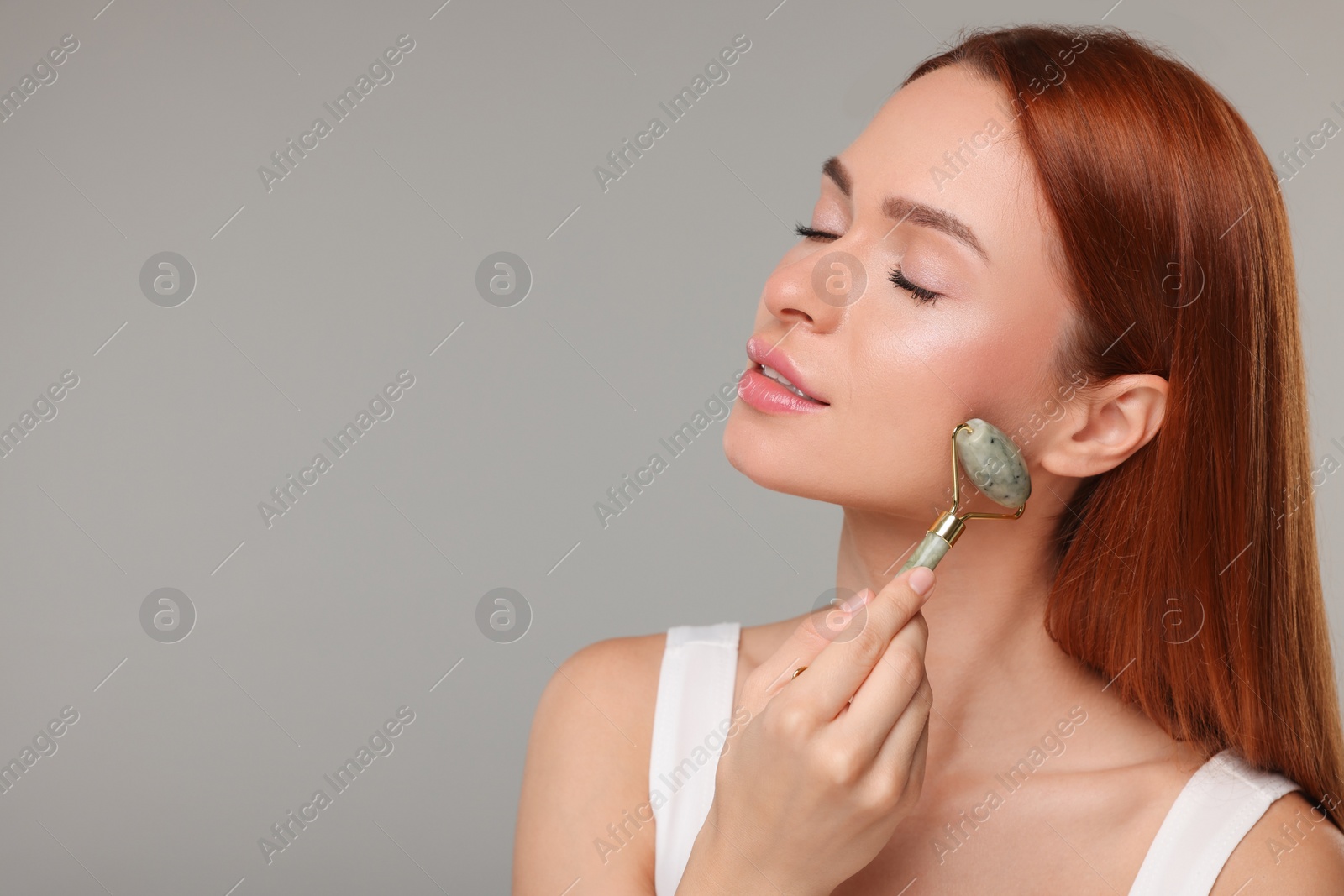 Photo of Young woman massaging her face with jade roller on grey background, space for text