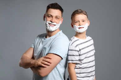 Happy dad and son with shaving foam on faces against grey background