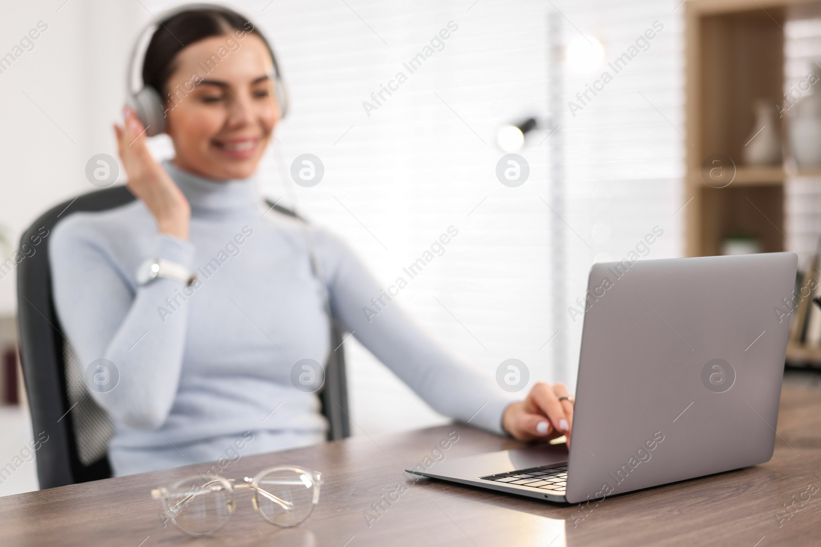 Photo of Young woman in headphones watching webinar at table in office, focus on laptop