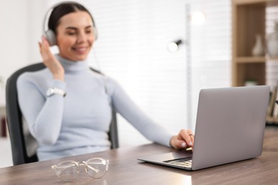 Young woman in headphones watching webinar at table in office, focus on laptop