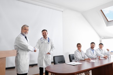 Photo of Doctors shaking hands near projection screen during medical conference in meeting room