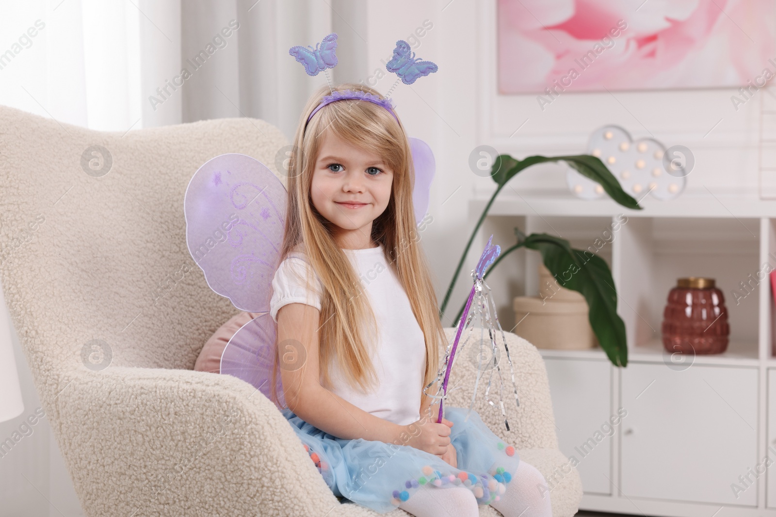 Photo of Cute little girl in fairy costume with violet wings and magic wand in armchair at home, space for text