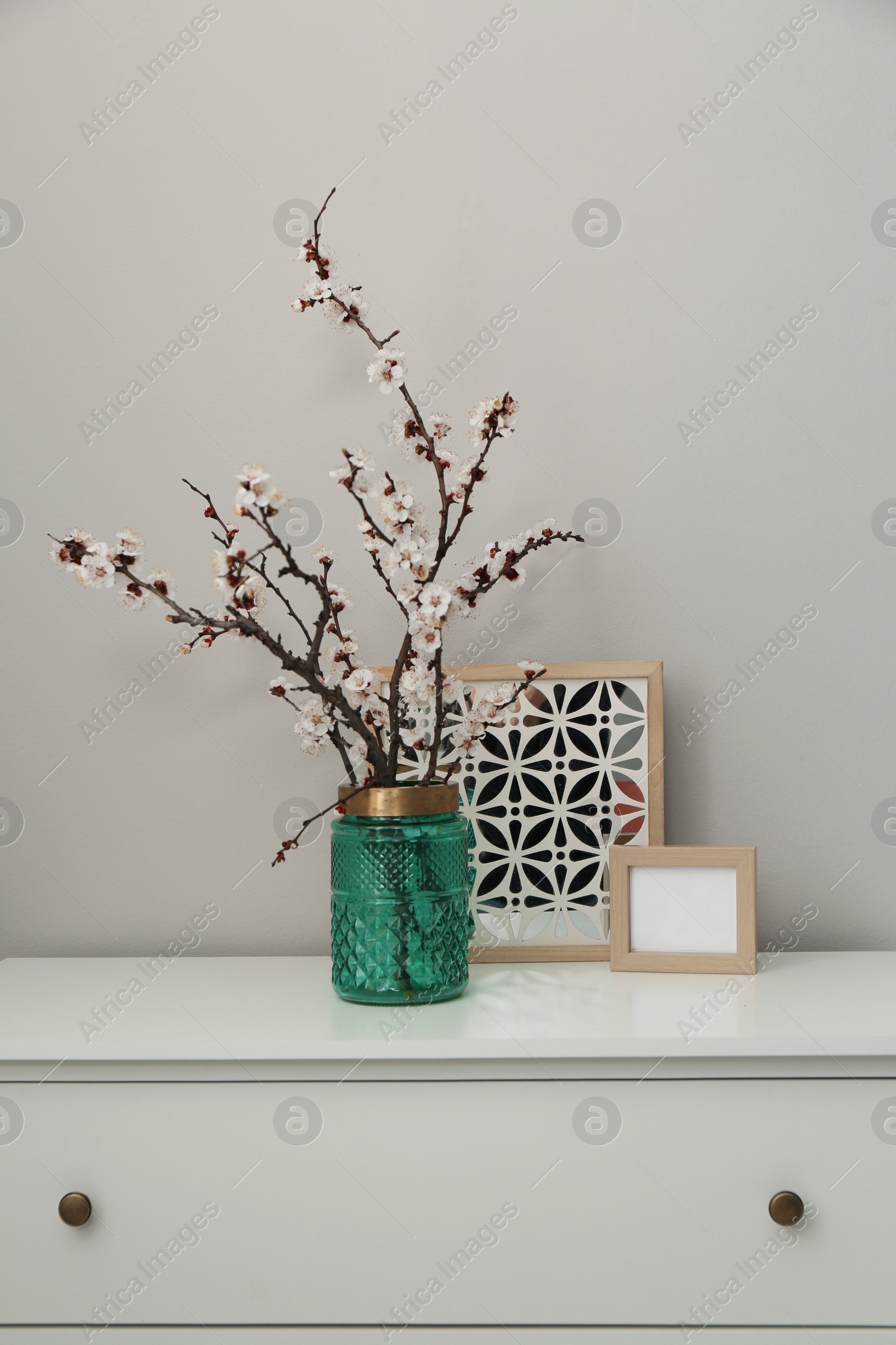 Photo of Flowering tree twigs in glass vase with decor on white chest of drawers near light wall