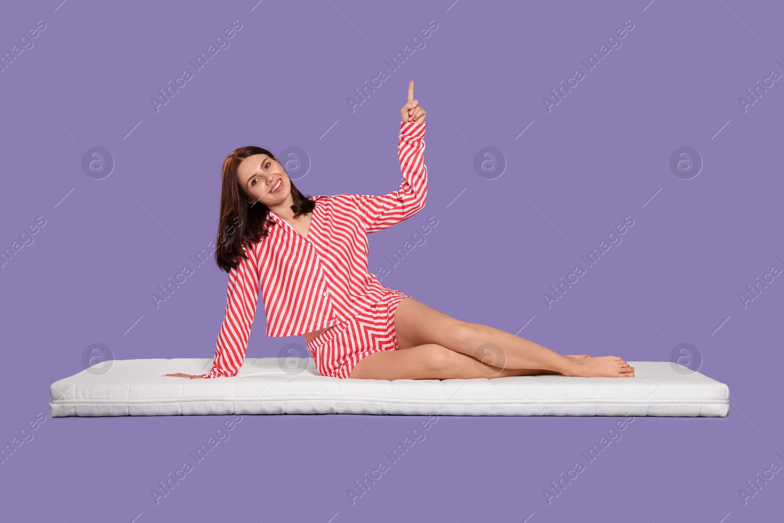 Photo of Young woman sitting on soft mattress and pointing upwards against light purple background