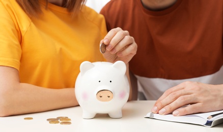 Couple putting coin into piggy bank at table, closeup. Saving money