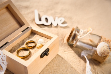 Wooden box with gold wedding rings on sandy beach, closeup