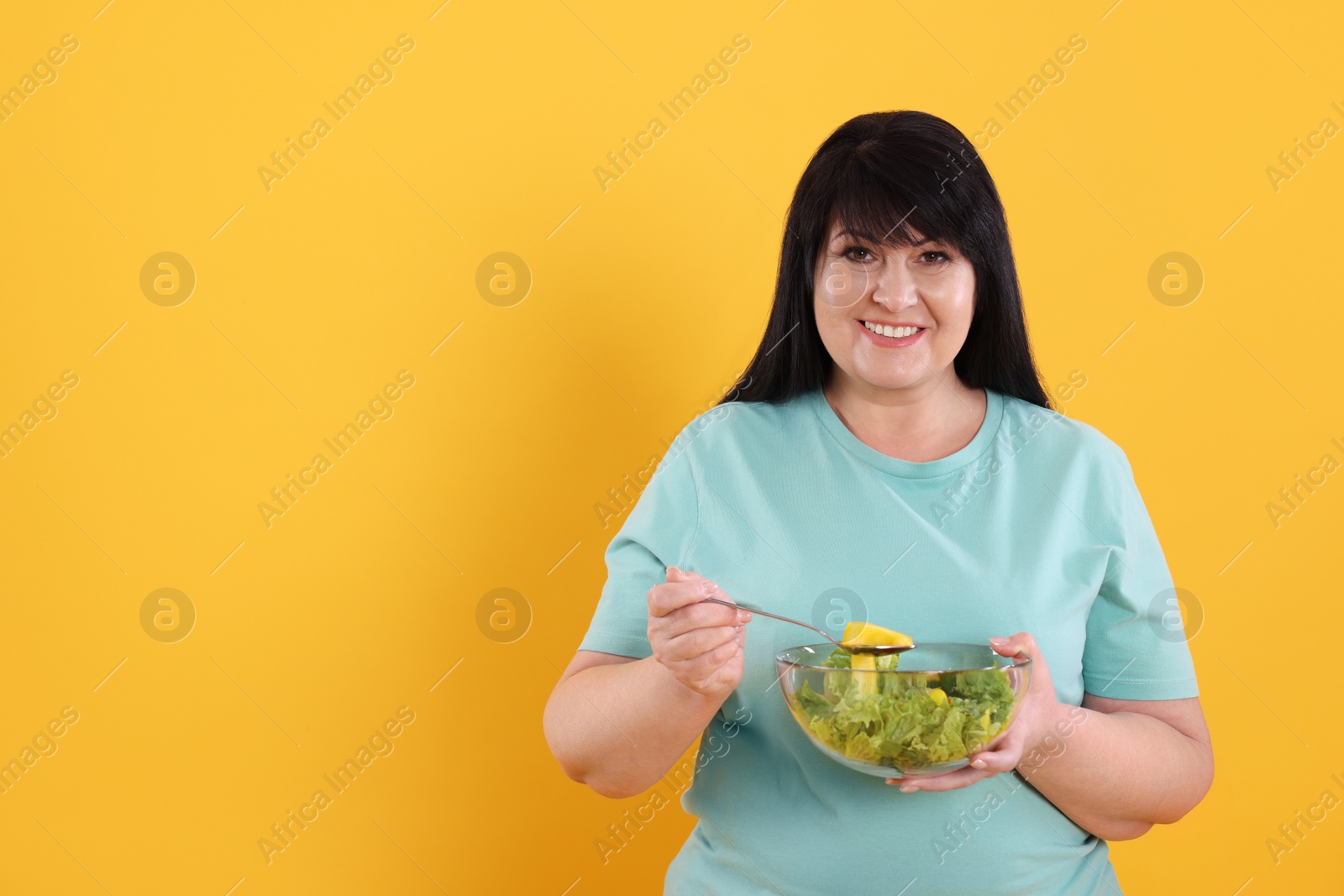 Photo of Beautiful overweight woman eating salad on yellow background, space for text. Healthy diet