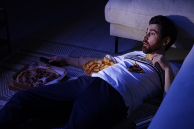 Photo of Man eating chips and pizza while watching TV in room at night. Bad habit
