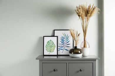 Photo of Reed's blossom in glass vases and pictures on grey cabinet indoors. Space for text