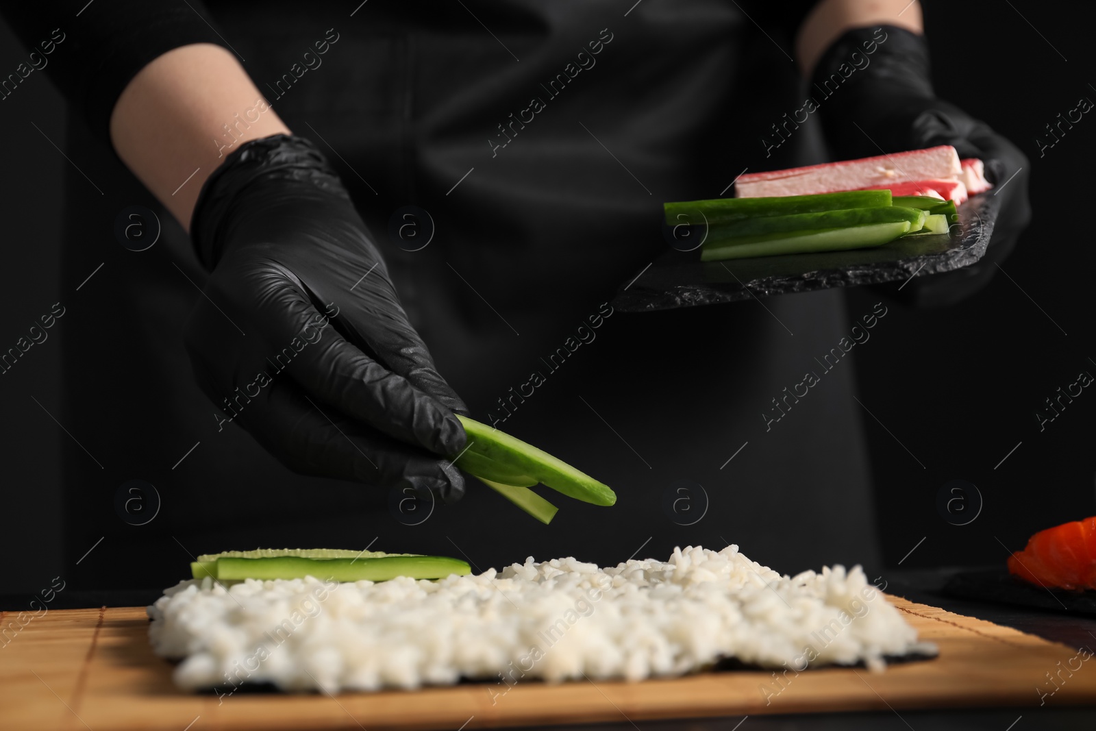 Photo of Chef in gloves putting cucumber onto unwrapped sushi roll at table, closeup