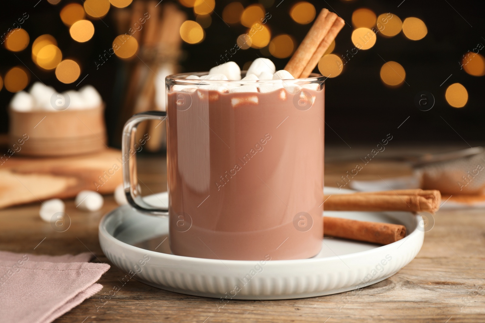 Photo of Glass cup of hot cocoa with aromatic cinnamon and marshmallows on wooden table against blurred lights