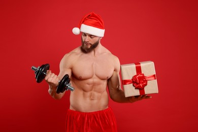 Muscular young man in Santa hat with dumbbell and Christmas gift box on red background