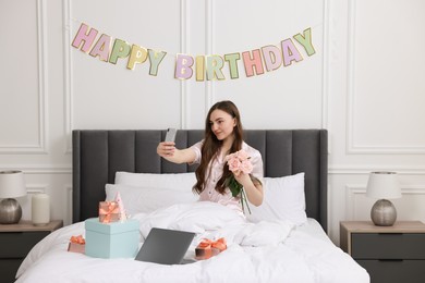 Photo of Beautiful young woman taking selfie with rose flowers, laptop and gift boxes on bed in room. Happy Birthday