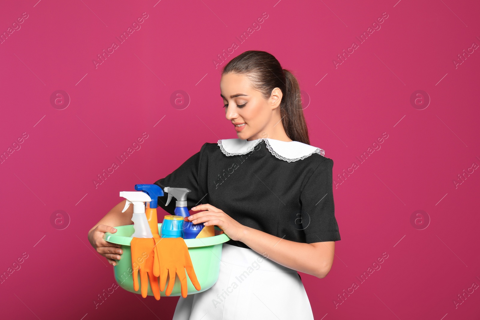 Photo of Young chambermaid holding plastic basin with detergents on color background