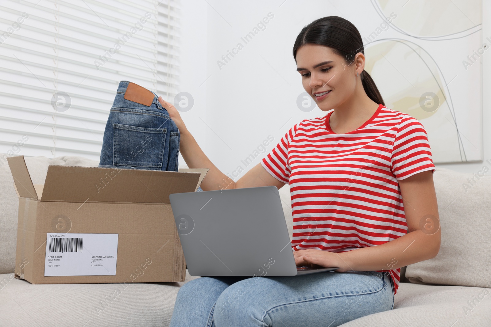 Photo of Young woman with just unpacked new jeans using laptop on sofa at home. Online shopping