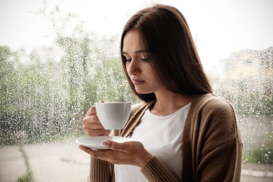 Photo of Thoughtful beautiful woman with cup of coffee near window indoors on rainy day