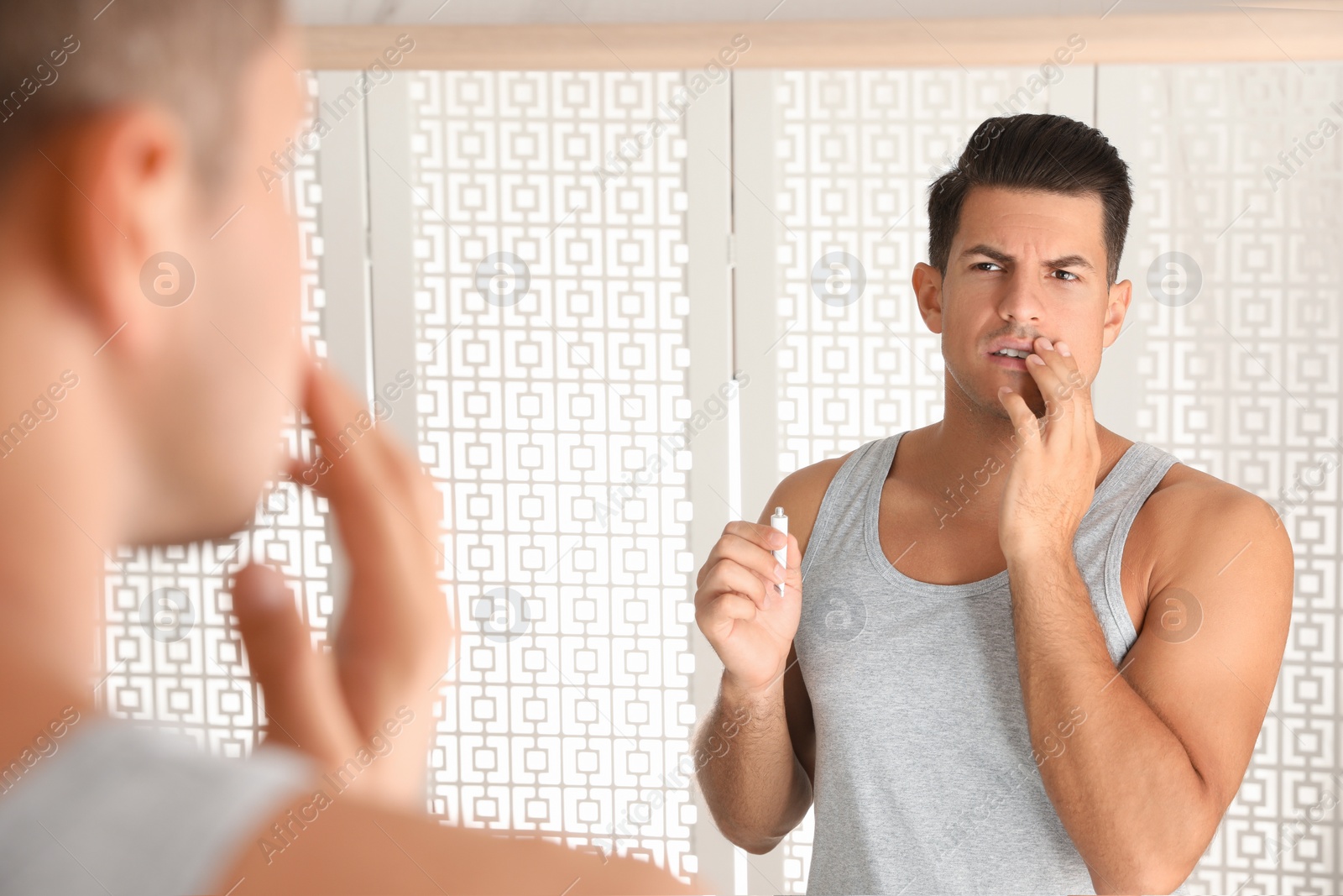 Photo of Emotional man with herpes applying cream on lips in front of mirror at home