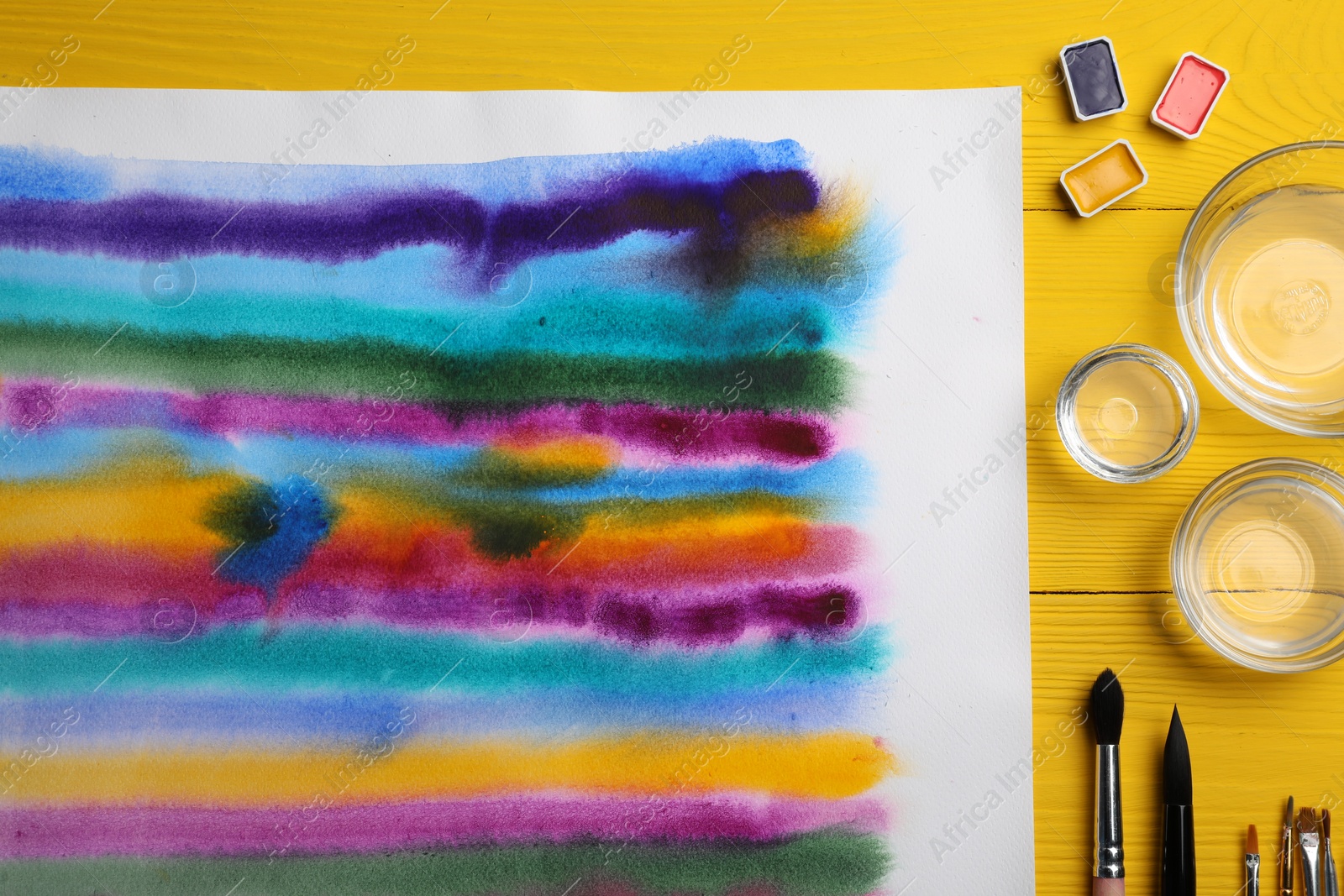 Photo of Bright abstract watercolor painting, paints, empty bowls and brushes on yellow wooden table, flat lay