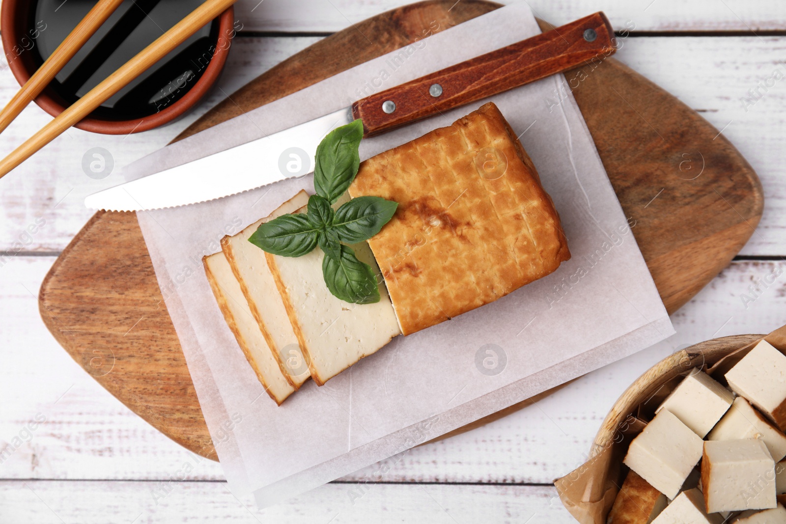 Photo of Board with smoked tofu, knife, basil and soy sauce on white wooden table, flat lay