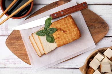 Photo of Board with smoked tofu, knife, basil and soy sauce on white wooden table, flat lay