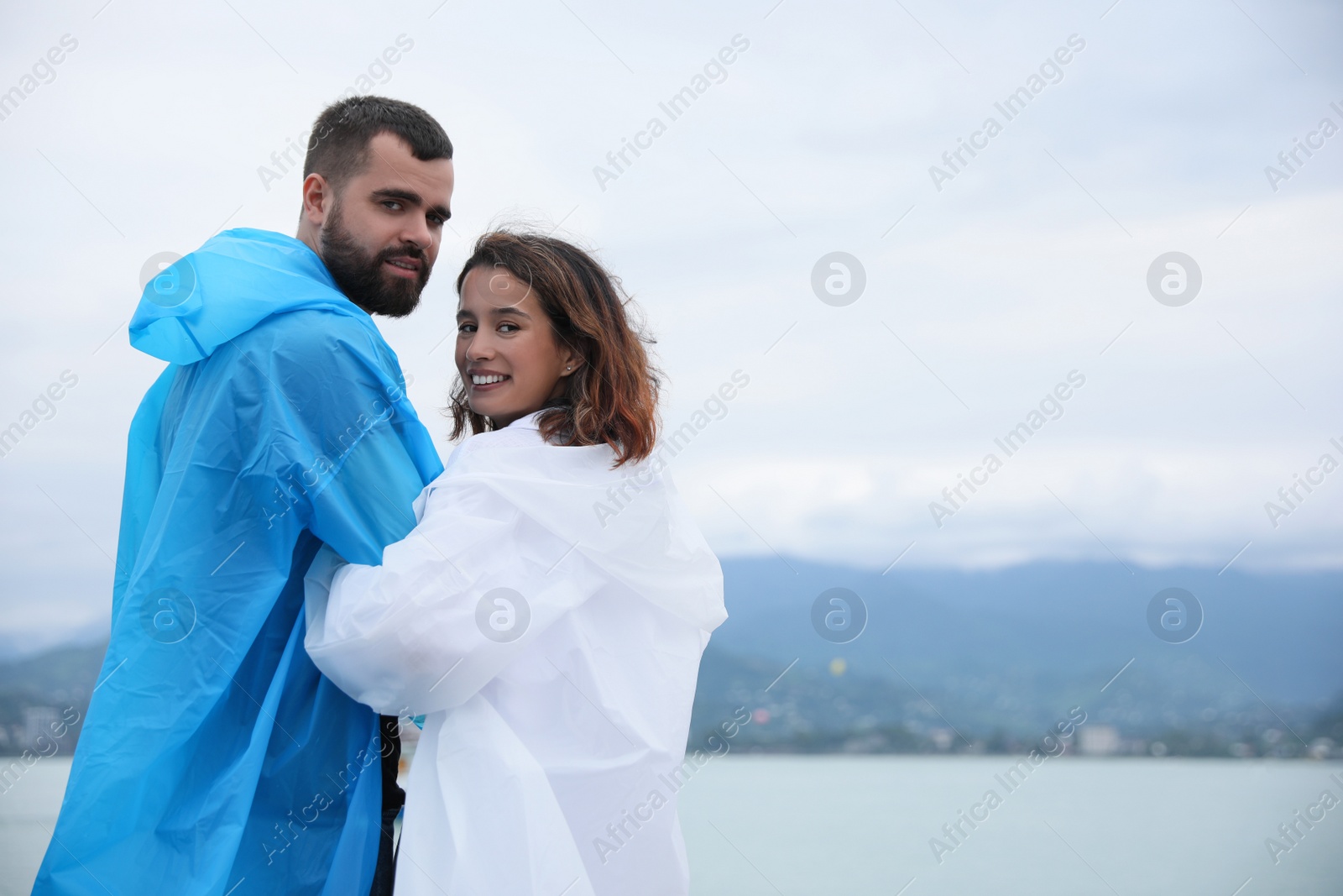 Photo of Young couple in raincoats enjoying time together under rain on beach, space for text