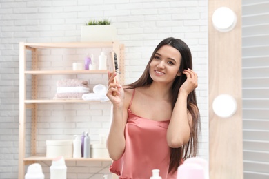 Beautiful young woman with hair brush looking into mirror in bathroom