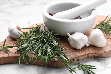 Photo of Sprigs of rosemary, mortar and garlic on white marble background, closeup