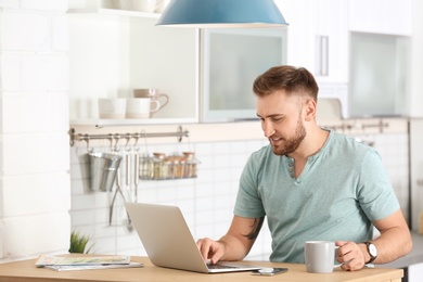 Photo of Young man using laptop at table in kitchen