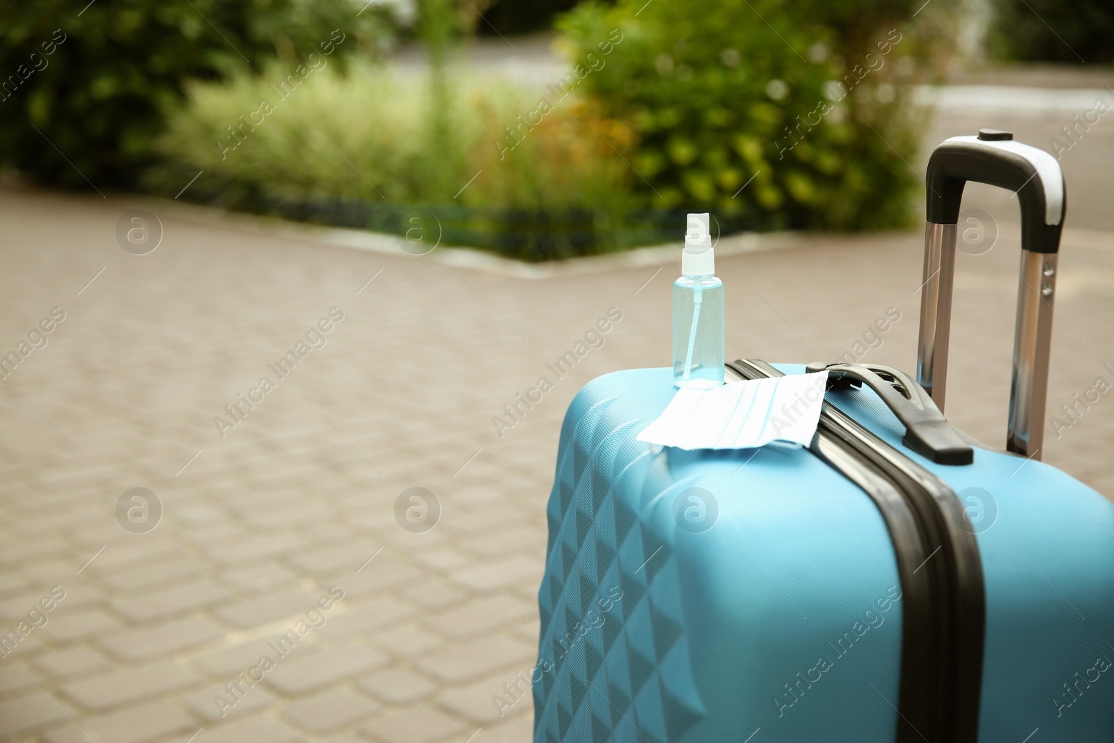 Photo of Stylish blue suitcase with protective mask and antiseptic spray outdoors, space for text. Travelling during coronavirus pandemic